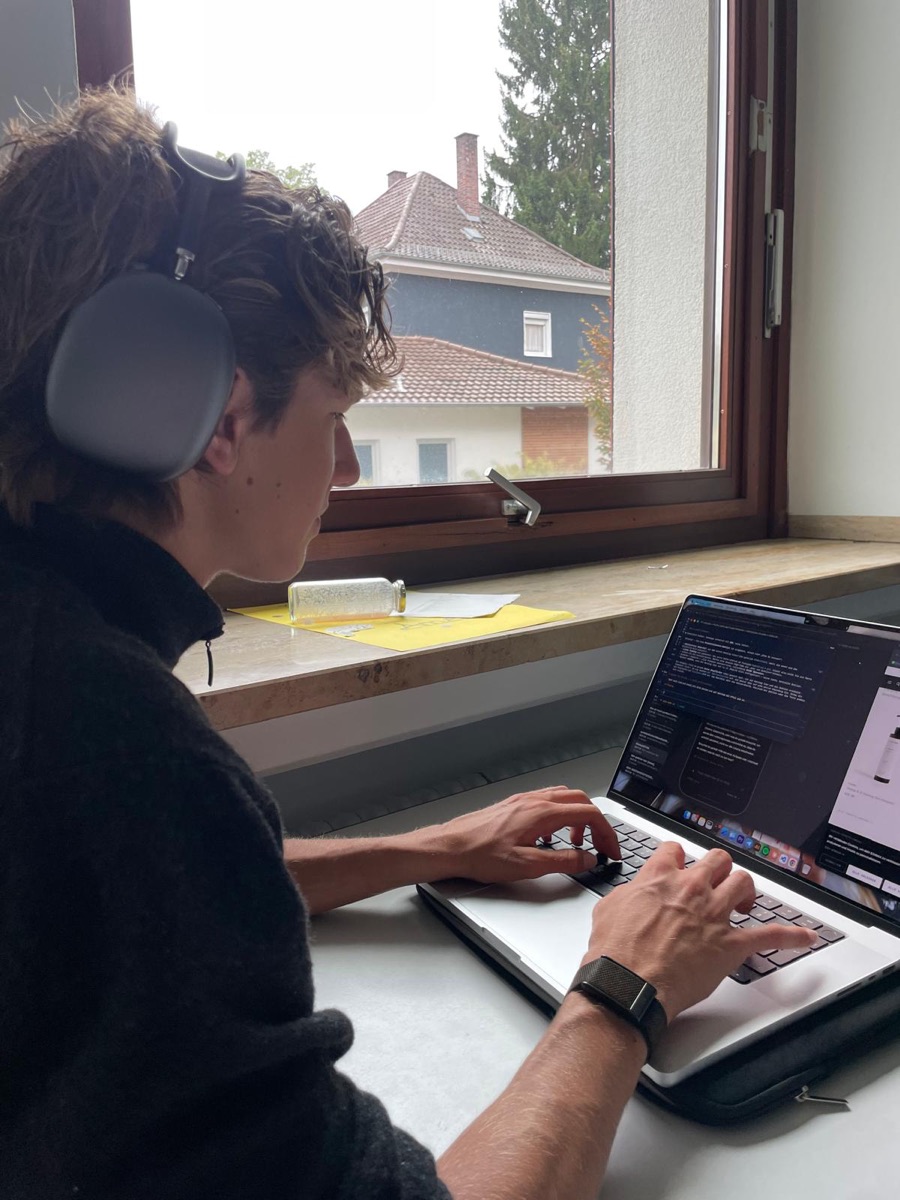 Elias Conrad im Profil bei der Arbeit am MacBook am Fenster, mit Kopfhörern